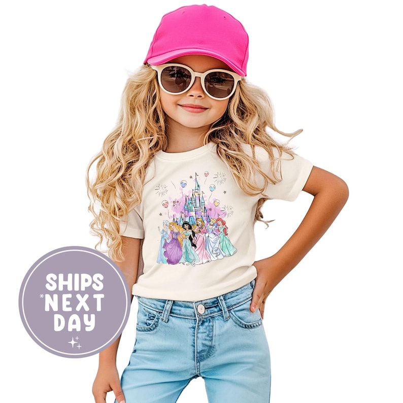 May include: A cream-colored t-shirt featuring a graphic of Disney princesses in front of a castle. The shirt is worn by a young person wearing a pink cap and sunglasses. The text "SHIPS NEXT DAY" is in a purple circle.