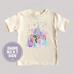 May include: Cream-colored t-shirt featuring a colorful illustration of several princesses in front of a castle with fireworks and balloons. The shirt has short sleeves and a round neckline. The text "SHIPS NEXT DAY" is on a circular graphic.