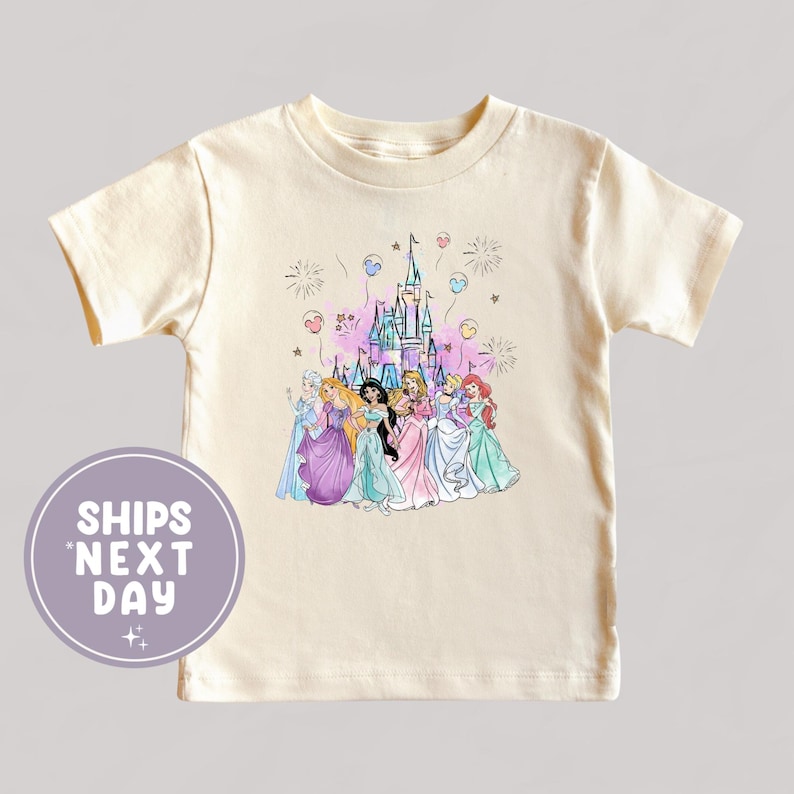 May include: Cream-colored t-shirt featuring a colorful illustration of several princesses in front of a castle with fireworks and balloons. The shirt has short sleeves and a round neckline. The text "SHIPS NEXT DAY" is on a circular graphic.