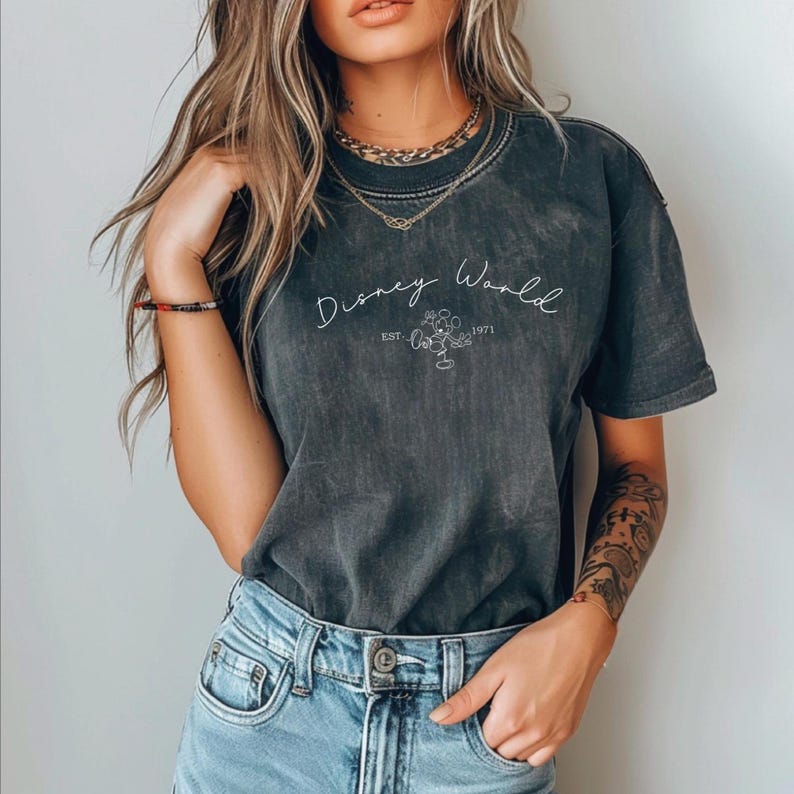 May include: Dark gray t-shirt with the words "Disney World" in cursive, along with a Mickey Mouse outline and the year "1971." The shirt has a vintage, washed look and is paired with blue jeans.