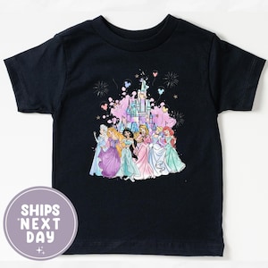 May include: A navy blue t-shirt featuring a colorful graphic of several princesses in front of a castle, with fireworks and balloons. The shirt has short sleeves and a round neckline. The text "SHIPS NEXT DAY" is printed on a round sticker.