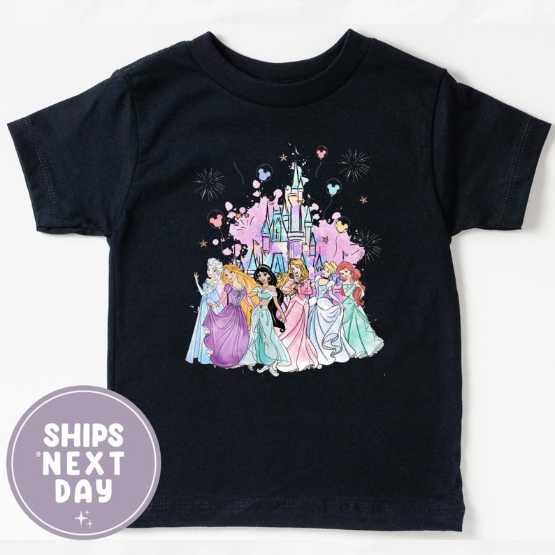 May include: A navy blue t-shirt featuring a colorful graphic of several princesses in front of a castle, with fireworks and balloons. The shirt has short sleeves and a round neckline. The text "SHIPS NEXT DAY" is printed on a round sticker.