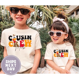 Pode incluir: Duas crianças vestindo camisetas creme com o texto "Cousin Crew" em preto, laranja e vermelho. As camisas têm os nomes "Mark" e "Olivia" impressos. Uma criança usa óculos de sol e um chapéu de palha.