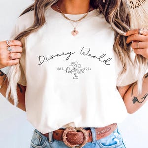 May include: Cream-colored t-shirt featuring the text "Disney World" in a cursive font, with "EST. 1971" below a line drawing of Mickey Mouse. The shirt is a casual wear item.