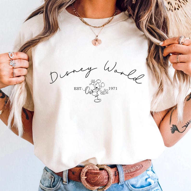 May include: Cream-colored t-shirt featuring the text "Disney World" in a cursive font, with "EST. 1971" below a line drawing of Mickey Mouse. The shirt is a casual wear item.