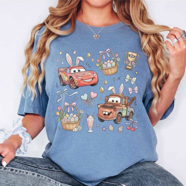 Może przedstawiać: Niebieska koszulka z kresk&oacute;wkowymi samochodami z kr&oacute;liczymi uszami, koszykami wielkanocnymi i innymi grafikami o tematyce wielkanocnej. Projekt zawiera trofeum, kokardy i koktajl mleczny. Koszulka jest w kolorze jasnoniebieskim.