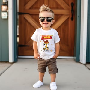 May include: A white t-shirt with a cowboy-themed graphic featuring the number "3" and the text "YOU'VE GOT A FRIEND IN ME THREE". The shirt is paired with brown shorts and white sneakers. The child is wearing black sunglasses.