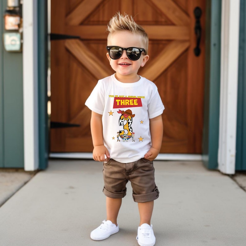 May include: A white t-shirt with a cowboy-themed graphic featuring the number "3" and the text "YOU'VE GOT A FRIEND IN ME THREE". The shirt is paired with brown shorts and white sneakers. The child is wearing black sunglasses.