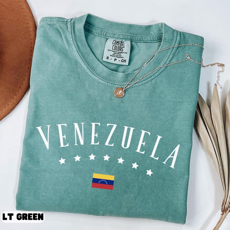 May include: A light green t-shirt with the word "VENEZUELA" in white arched text, with five white stars below. The Venezuelan flag is displayed below the stars. A gold necklace with a pendant rests on the shirt. The shirt is labeled "LT GREEN".