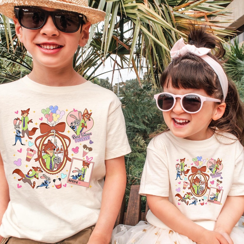 Może przedstawiać: Dwoje dzieci w kremowych koszulkach z kolorowym wzorem z kresk&oacute;wek. Wz&oacute;r przedstawia postać lisa w zielonej koszuli, kr&oacute;lika, balony oraz napisy "Best Friends Forever" i "Bestie".