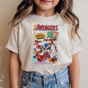 May include: Cream-colored t-shirt featuring a comic book-style graphic with the word "AVENGERS" in red. The graphic includes Disney characters like Mickey Mouse, Donald Duck, and Iron Man in action poses. The shirt has short sleeves.