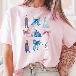 May include: A light pink t-shirt featuring various illustrations of characters and elements from a popular animated movie. The design includes a castle, bows, and characters in shades of blue, pink, and white.