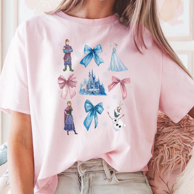 May include: A light pink t-shirt featuring various illustrations of characters and elements from a popular animated movie. The design includes a castle, bows, and characters in shades of blue, pink, and white.