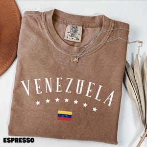 May include: Espresso-colored t-shirt with the word "VENEZUELA" in white, along with white stars and the Venezuelan flag. A silver necklace with a pendant rests on the shirt. The shirt is a Comfort Colors brand.