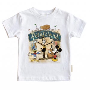 May include: White t-shirt featuring a colorful graphic of Disney characters Donald Duck, Goofy, and Mickey Mouse. The graphic includes the text "Potatoland" above the characters. The design is set against a whimsical, vintage-style background.