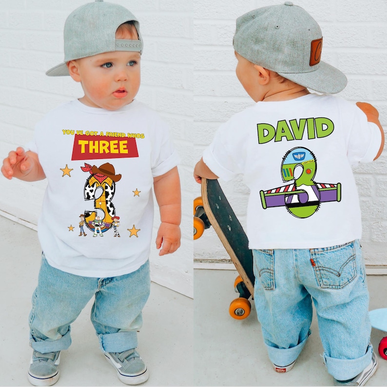 May include: White t-shirt with a graphic design featuring the number "3" in a cowboy-themed design, with the text "YOU'VE GOT A FRIEND IN ME" and "THREE". The back of the shirt has the name "DAVID" and a Buzz Lightyear-themed "3".