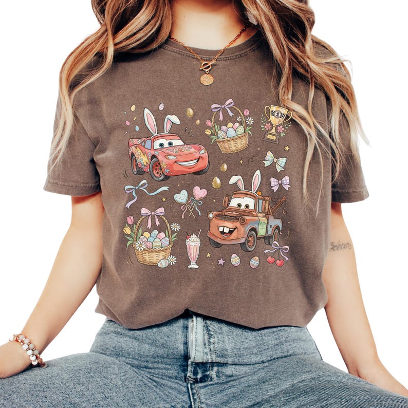 Może przedstawiać: Brązowa koszulka z kolorową grafiką wielkanocną. Projekt zawiera kresk&oacute;wkowe samochody z kr&oacute;liczymi uszami, koszyki wielkanocne, trofeum, kokardy i pisanki. Koszulka ma klasyczny okrągły dekolt i kr&oacute;tkie rękawy.