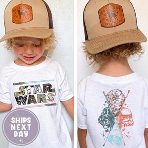 May include: A white Star Wars t-shirt with the Star Wars logo and lightsabers. The back of the shirt features Darth Vader, a Stormtrooper, and the Death Star. The child is wearing a brown and tan trucker hat with a logo.