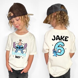 K&ouml;nnte beinhalten: Cremefarbenes T-Shirt mit einer Cartoonfigur und dem Text "Birthday Boy" auf der Vorderseite. Auf der R&uuml;ckseite stehen der Name "Jake" und die Zahl "6" mit derselben Cartoonfigur. Das T-Shirt wird von einem Kind getragen.
