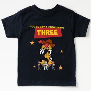 May include: Navy blue t-shirt with a graphic featuring the number "3" designed as a cowboy with a hat and bandana. The shirt includes the text "YOU'VE GOT A FRIEND WHOS THREE" and images of cartoon characters and stars.