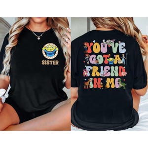 May include: Black t-shirt featuring a cartoon alien with the word "SISTER" on the front. The back of the shirt displays the phrase "YOU'VE GOT A FRIEND IN ME" with various cartoon characters. The design is in a retro style.