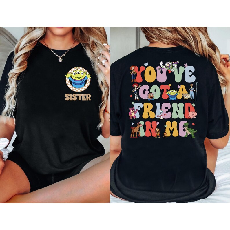 May include: Black t-shirt featuring a cartoon alien with the word "SISTER" on the front. The back of the shirt displays the phrase "YOU'VE GOT A FRIEND IN ME" with various cartoon characters. The design is in a retro style.