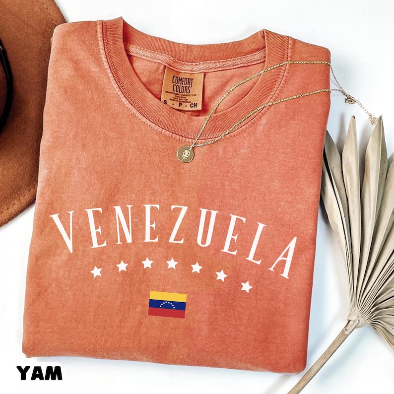 May include: A coral-colored t-shirt with the word "VENEZUELA" in white block letters, along with a Venezuelan flag graphic and stars. A gold necklace with a pendant rests on the shirt. The shirt has a Comfort Colors tag.