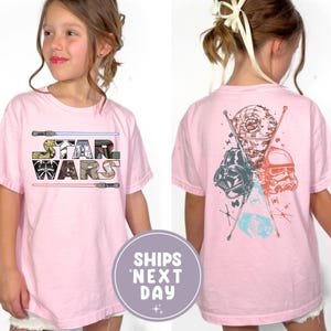 May include: A light pink t-shirt featuring the Star Wars logo on the front and character graphics on the back. The front design includes the words "STAR WARS" in a multi-colored font with lightsabers. The back features Darth Vader, a Stormtrooper, and the Death Star.
