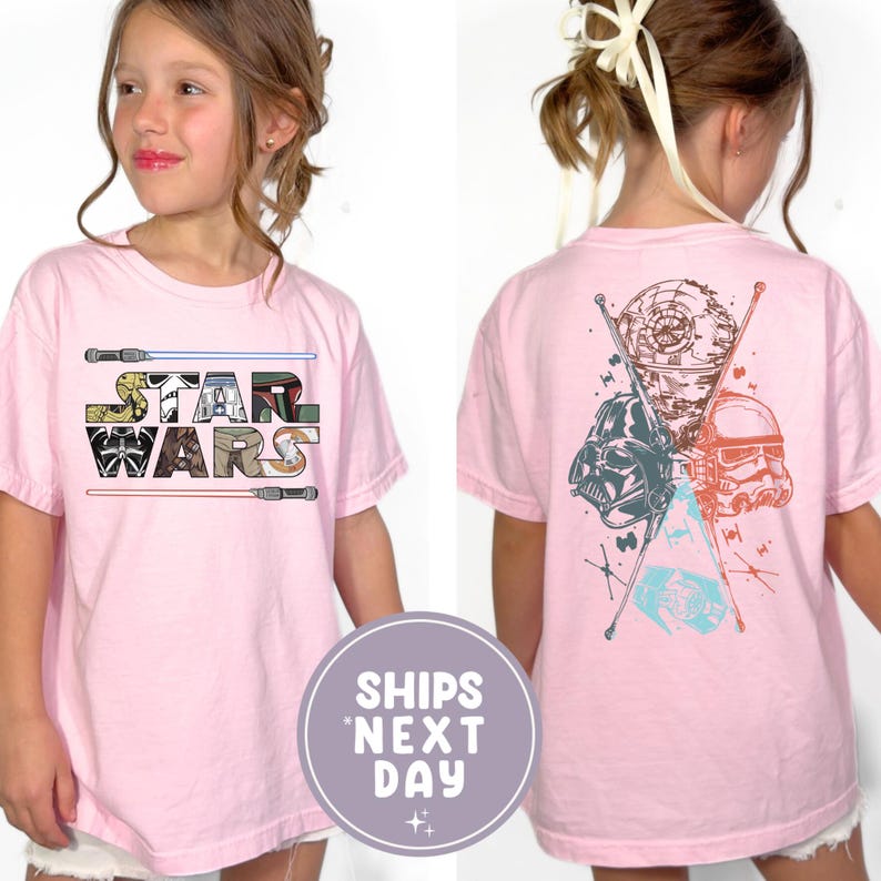 May include: A light pink t-shirt featuring the Star Wars logo on the front and character graphics on the back. The front design includes the words "STAR WARS" in a multi-colored font with lightsabers. The back features Darth Vader, a Stormtrooper, and the Death Star.