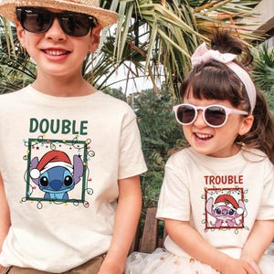 May include: Two children wearing matching cream-colored t-shirts with Christmas-themed Stitch graphics. One shirt says "DOUBLE" and the other says "TROUBLE". Both shirts feature Stitch wearing a Santa hat and surrounded by Christmas lights.