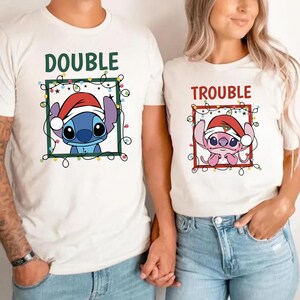 May include: White t-shirts with matching graphics. One shirt says "DOUBLE" with a blue cartoon character wearing a Santa hat. The other shirt says "TROUBLE" with a pink cartoon character wearing a Santa hat. Both characters are framed with green and red borders and Christmas lights.
