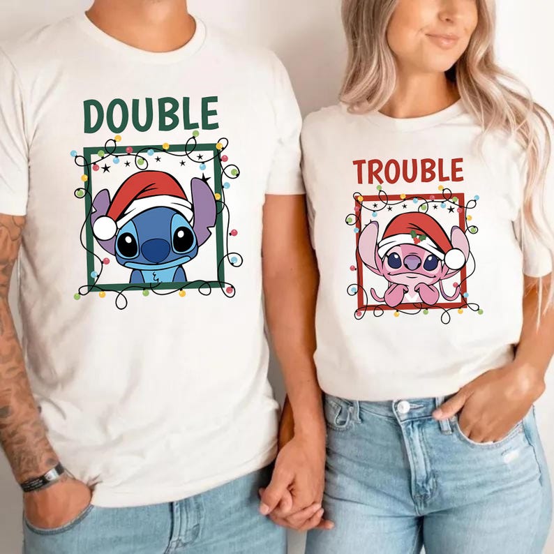 May include: White t-shirts with matching graphics. One shirt says "DOUBLE" with a blue cartoon character wearing a Santa hat. The other shirt says "TROUBLE" with a pink cartoon character wearing a Santa hat. Both characters are framed with green and red borders and Christmas lights.