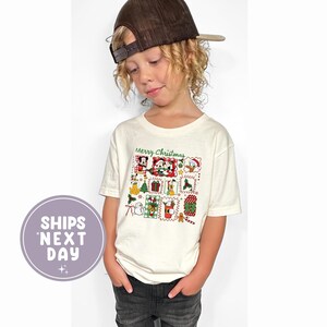 May include: Cream-coloured short-sleeved t-shirt with a festive Christmas graphic featuring Mickey Mouse and friends. The words "Merry Christmas" are printed above the graphic. The shirt is worn by a child, who is also wearing a brown baseball cap.