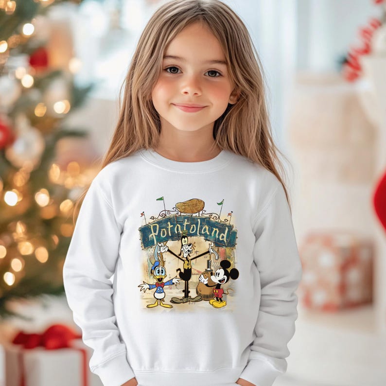 May include: A white sweatshirt featuring a colorful graphic with the text "Potatoland" above cartoon characters Donald Duck, Goofy, and Mickey Mouse. The sweatshirt is worn by a young girl.