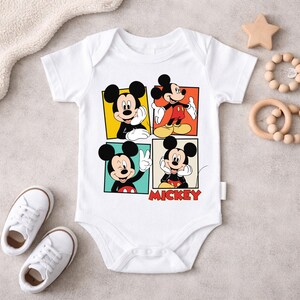 May include: White baby bodysuit featuring four panels with different poses of Mickey Mouse. The word "Mickey" is printed in red at the bottom. The bodysuit is surrounded by baby shoes, a wooden star, and a teething ring.