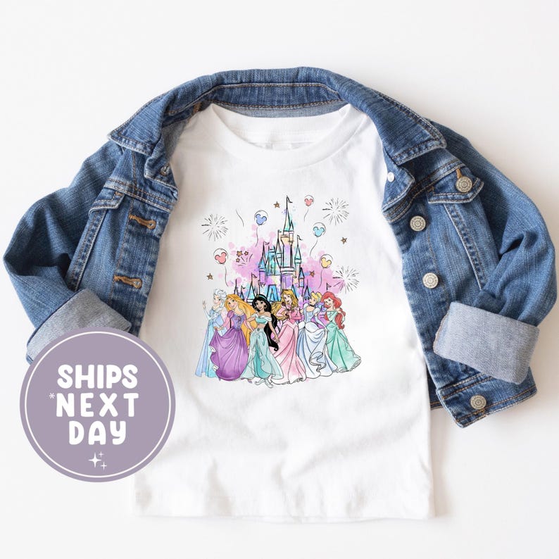 May include: White t-shirt featuring a watercolor illustration of several princesses in colorful gowns in front of a castle. The shirt is overlaid by a blue denim jacket. The text "SHIPS NEXT DAY" is in a purple circle.