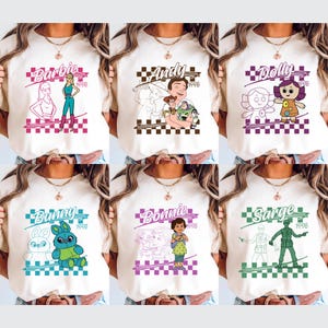 Op de afbeelding: Zes cr&egrave;mekleurige t-shirts met verschillende grafische ontwerpen. Elk shirt heeft een personagenaam, zoals "Barbie" of "Andy", met een bijbehorende illustratie en het jaartal "1996" in een geblokte kader. De ontwerpen zijn in verschillende kleuren.