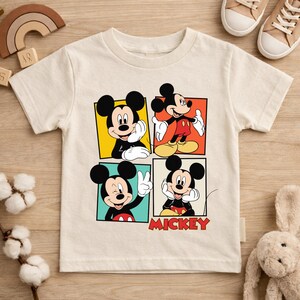 May include: Off-white short-sleeved t-shirt featuring a colorful Mickey Mouse graphic. The graphic is divided into four squares, each showcasing a different pose of Mickey. The word "MICKEY" is printed in red at the bottom.
