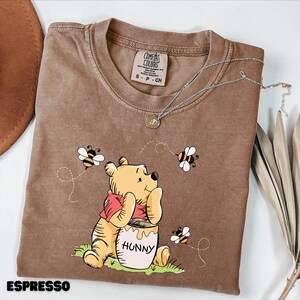 May include: Espresso-colored t-shirt featuring a graphic of Winnie the Pooh with a honey pot and bees. The shirt has a crew neck and short sleeves. A silver necklace with a pendant is draped across the shirt. The text "ESPRESSO" is at the bottom.
