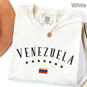 May include: A white t-shirt with the word "VENEZUELA" in black, arched above a row of black stars and a small Venezuelan flag. A gold necklace with a pendant rests on the shirt. The shirt is labeled "COMFORT COLORS".