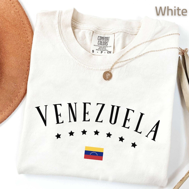 May include: A white t-shirt with the word "VENEZUELA" in black, arched above a row of black stars and a small Venezuelan flag. A gold necklace with a pendant rests on the shirt. The shirt is labeled "COMFORT COLORS".
