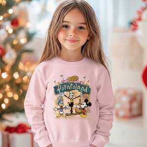 May include: A light pink sweatshirt featuring a cartoon graphic of Donald Duck, Goofy, and Mickey Mouse under a sign that reads "Potatoland". The sweatshirt is worn by a young girl with long brown hair.