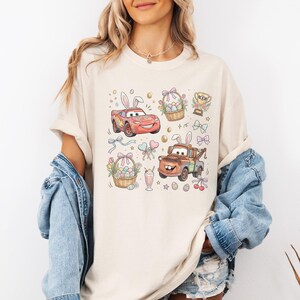 Może przedstawiać: Kremowy t-shirt z kolorową grafiką o tematyce wielkanocnej. Projekt zawiera kresk&oacute;wkowe samochody z kr&oacute;liczymi uszami, koszyki wielkanocne z jajkami, trofeum, kokardy i inne świąteczne elementy. Koszulka ma kr&oacute;tkie rękawy i luźny kr&oacute;j.