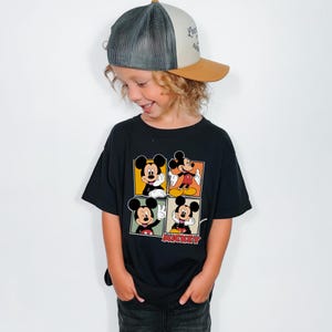 May include: A black t-shirt featuring four panels with different images of Mickey Mouse. The shirt has a crew neck and short sleeves. The child is also wearing a trucker hat with a gray mesh back and a brown brim.