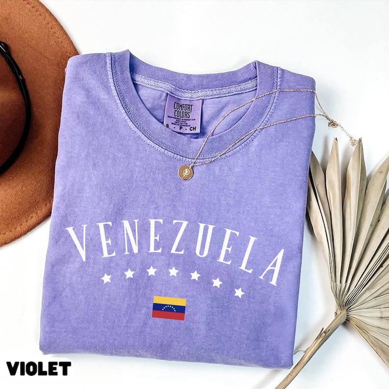 May include: A violet Comfort Colors t-shirt with the word "VENEZUELA" in white, along with five white stars and the Venezuelan flag. A gold necklace with a pendant rests on the shirt. A brown hat and dried plant are in the background.