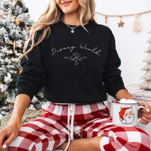 May include: Black crew neck sweatshirt with the text "Disney World" in a script font, along with a Mickey Mouse graphic and the year "1971". The person is wearing red and white plaid pajama pants and holding a mug with a Santa Claus design.