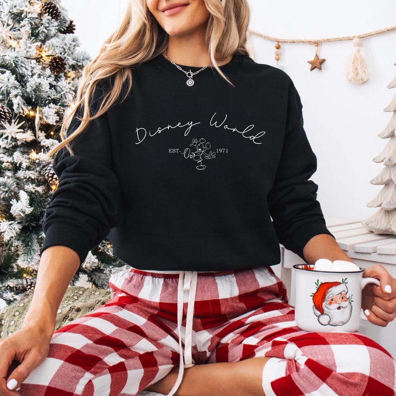 May include: Black crew neck sweatshirt with the text "Disney World" in a script font, along with a Mickey Mouse graphic and the year "1971". The person is wearing red and white plaid pajama pants and holding a mug with a Santa Claus design.