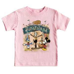 May include: A light pink toddler t-shirt featuring a cartoon graphic of Mickey Mouse, Donald Duck, and Goofy in front of a sign that reads "Potatoland". The characters are in a vintage style.
