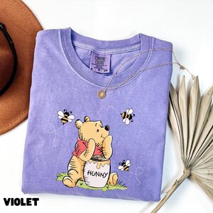 May include: Violet Comfort Colors t-shirt featuring a graphic of Winnie the Pooh with a honey pot and bees. The shirt has a crew neck and short sleeves. A gold necklace with a pendant is draped across the shirt.