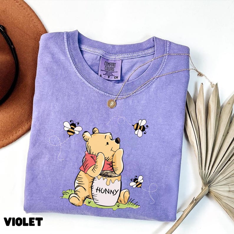 May include: Violet Comfort Colors t-shirt featuring a graphic of Winnie the Pooh with a honey pot and bees. The shirt has a crew neck and short sleeves. A gold necklace with a pendant is draped across the shirt.